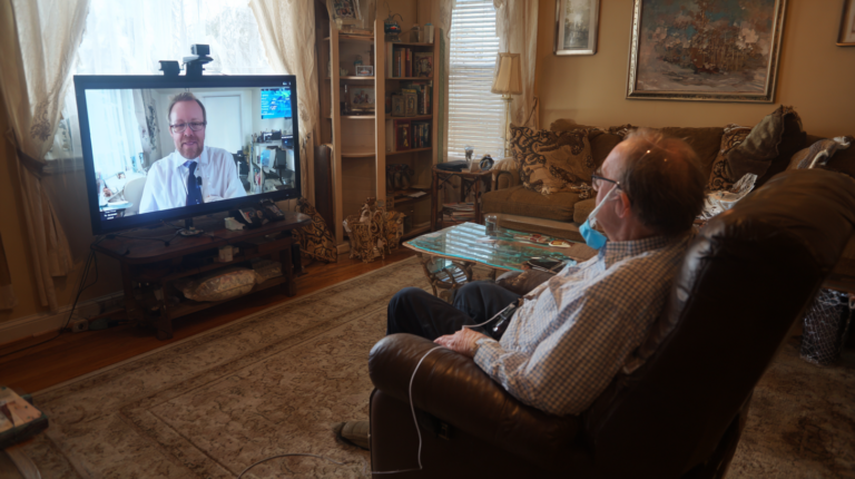 An older man with an IV on a recliner is having a telemedicine conference with a doctor on a tv.