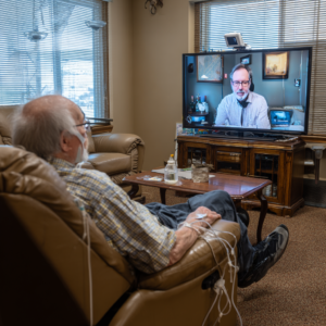 An older man is on a video call with a man in a tie on a tv.